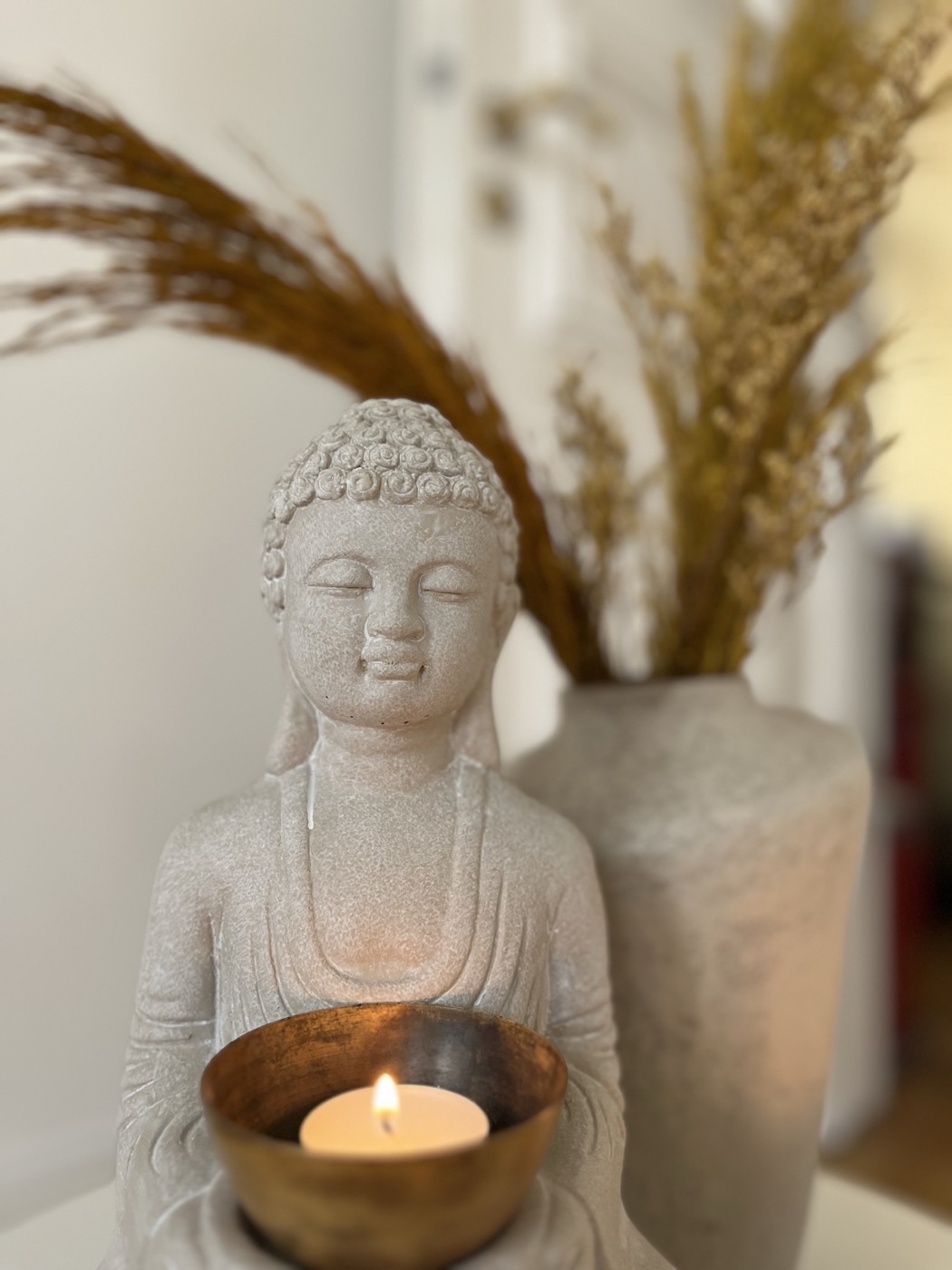 Ruhige Buddha-Figur hält eine kleine Messingschale mit brennendem Teelicht, getrocknetes goldenes Pampasgras im Hintergrund